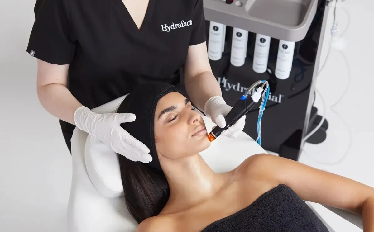 Eine Kundin von Hydrafacial erhält eine Hydrafacial Behandlung, von der Kosmetikerin die das Hydrafacial-Gerät benutzt. Sie trägt ein dunkles Tuch auf ihrem Kopf und wird mit dem original Hydrafacial-Aufsatz behandelt. Die behandelnde Kosmetikerin trägt dabei weiße Handschuhe